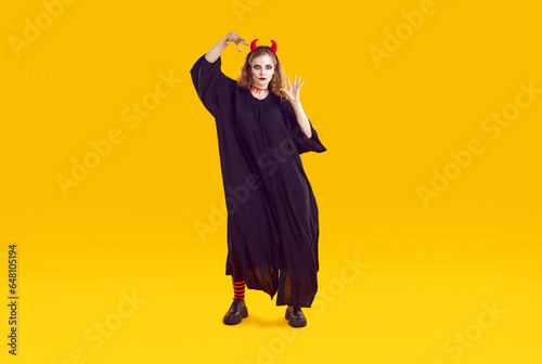 Woman in Halloween costume is scaring you with creepy serious look and waving her hands. Girl in long black dress, with red horns and bloody cut on throat is isolated on orange background. Full length © Studio Romantic