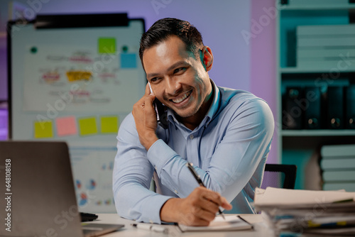 Asian man talking on the phone in the office of a startup company, company employees working overtime, late night work, overtime of a busy and hard working company employee. Overtime concept.