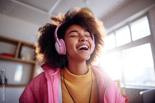 Happy funny gen z hipster African American teen girl wearing headphones dancing at home, listening music on mobile phone, having fun feeling funky moving in living room, authentic shot