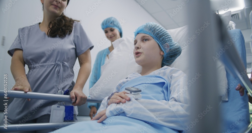 Girl lies on gurney, speaks with nurse and mother, smiles and shows pulse oximeter to medic. Doctor and nurse transport young patient to hospital ward down clinic corridor. Medical staff at work. Girl lies on gurney, speaks with nurse and mother, smiles and shows pulse oximeter to medic. Doctor and nurse transport young patient to hospital ward down clinic corridor. Medical staff at work.
