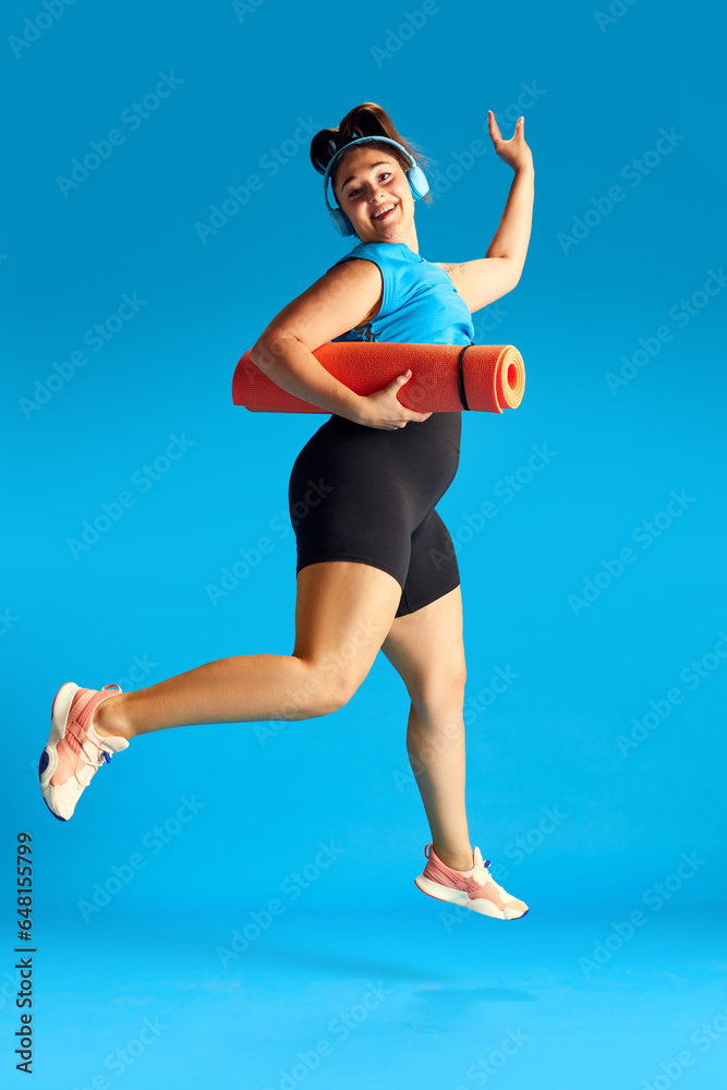Excited and positive woman in sportswear, in headphones running with fitness matt to training against blue background. Concept of sport, healthy lifestyle, human emotions, fashion, ad