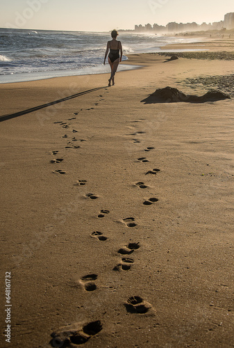 Mature woman walks away along the beach at sunset next to the sea, leaving a trace of footprints in the sand, in the background the mist and buildings, in Punta del Este, Uruguay.. photo