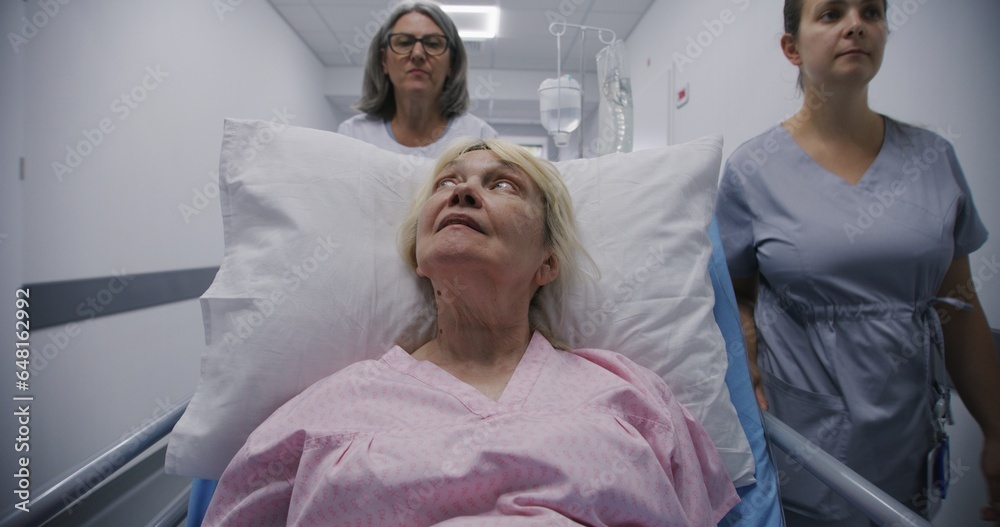 Fototapeta premium Mature female doctor with young nurse brings senior patient to medical center. Old woman lies on gurney with drip and talks with medics about surgery. Medical staff at work in modern hospital.