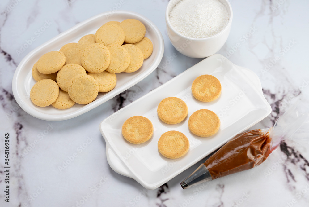ingredients and mise en place for the preparation of cornstarch alfajores, filling the dulce de leche filling on top and grated coconut on the outside on white plates. ingredients and mise en place for the preparation of cornstarch alfajores, filling the dulce de leche filling on top and grated coconut on the outside on white plates.