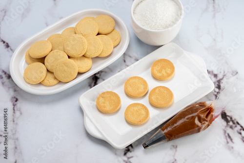 ingredients and mise en place for the preparation of cornstarch alfajores, filling the dulce de leche filling on top and grated coconut on the outside on white plates.