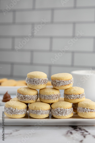 Homemade cornstarch alfajores filled with dulce de leche with coconut and cup of tea on plates and white background photo