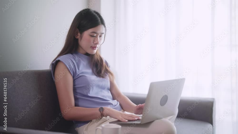 Pretty Asian woman surfing social media or working on laptop while lying on the sofa when relax in living room at home., Concept of activity in living room. Pretty Asian woman surfing social media or working on laptop while lying on the sofa when relax in living room at home., Concept of activity in living room.