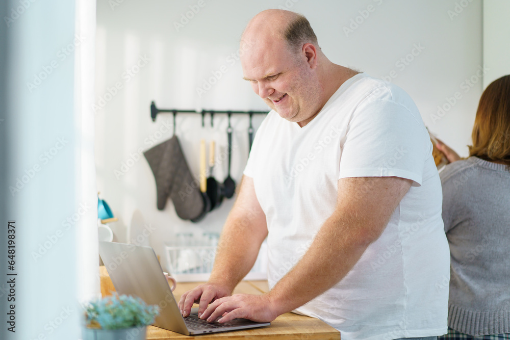 Caucasian - white oversize obese man checking an emails and messages on laptop - notebook computer on the counter top in kitchen between drinks a cup of coffee in morning. Caucasian - white oversize obese man checking an emails and messages on laptop - notebook computer on the counter top in kitchen between drinks a cup of coffee in morning.