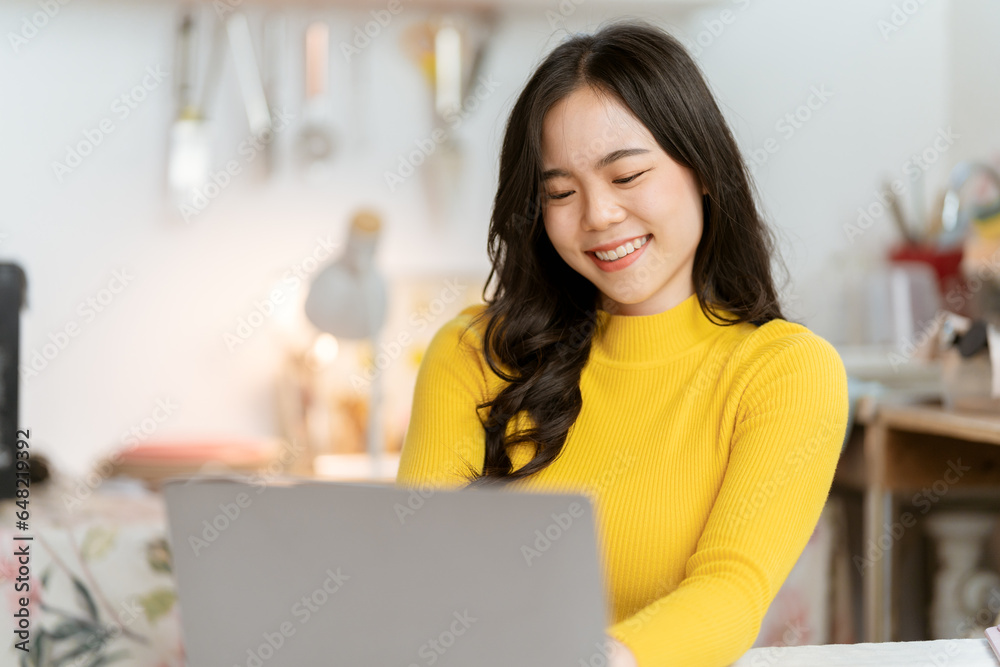 Beautiful Asian woman smiling happily relaxing using technology of laptop computer Take notes Drink a relaxing hot drink while sitting on the table in your room at home.