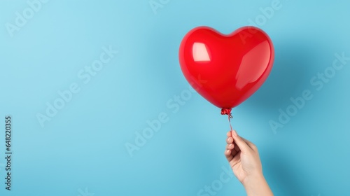 Female hand holding red heart shaped balloon on light blue background with copy space. Valentine's Day or Happy Birthday celebration concept.