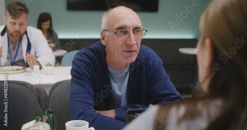 Elderly patient sits in clinic cafe with female doctor. Healthcare specialist discusses medical diagnostic results with man. Medic eats dinner in the background. Hospital or medical center cafeteria.