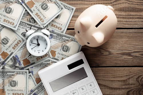 Many dollar bills with alarm clock and calculator, piggy bank on wooden table. Business concept. Time is money