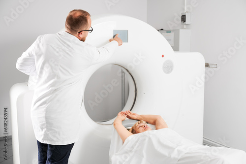 Medical computed tomography or MRI scanner. Back view of male radiologist presses MRI button to examine female patient.
