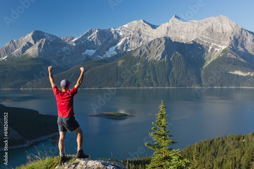 A Man Stands With Arms Raised On A Rock Overlooking A Lake And The Rocky Mountains; Kananaskis, Alberta, Canada photo