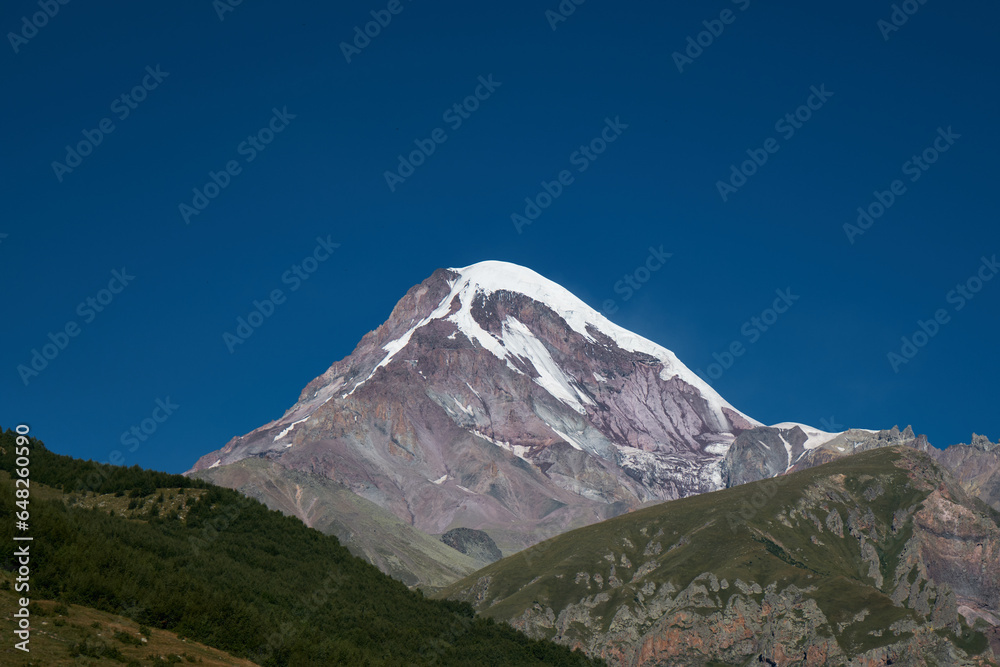 Nature of the North Caucasus. The snowy peak of Mount Kazbek. View from the peak in the village of Stepantsminda, Kazbegi. Blue cloudless sky. Trip to Georgia.