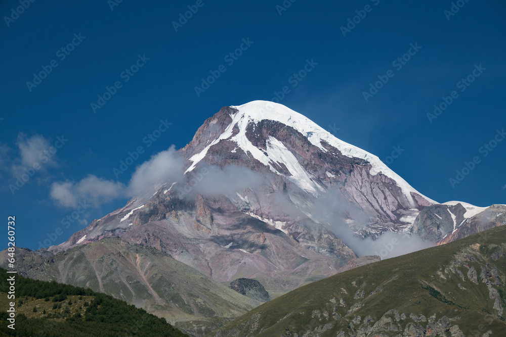 Nature of the North Caucasus. The snowy peak of Mount Kazbek. View from the peak in the village of Stepantsminda, Kazbegi. Blue cloudy sky. Trip to Georgia. A popular point of view for tourists.