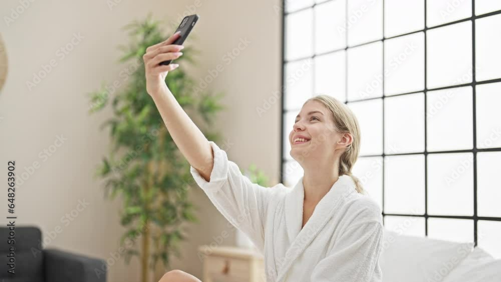 Young blonde woman wearing bathrobe sitting on bed make selfie by smartphone at bedroom