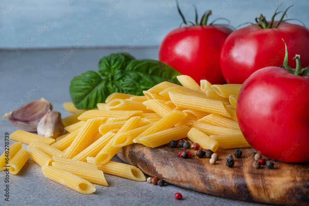 Blurred image of penne pasta, tomatoes, basil on a wooden cutting board on a gray background. Blurred image of penne pasta, tomatoes, basil on a wooden cutting board on a gray background.