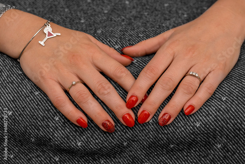 Hands of a girl resting on a uniform gray towel. Nails with freshly applied shiny red nail polish. Ring with very brilliant trilogy diamond and bracelet with cocktail symbol.