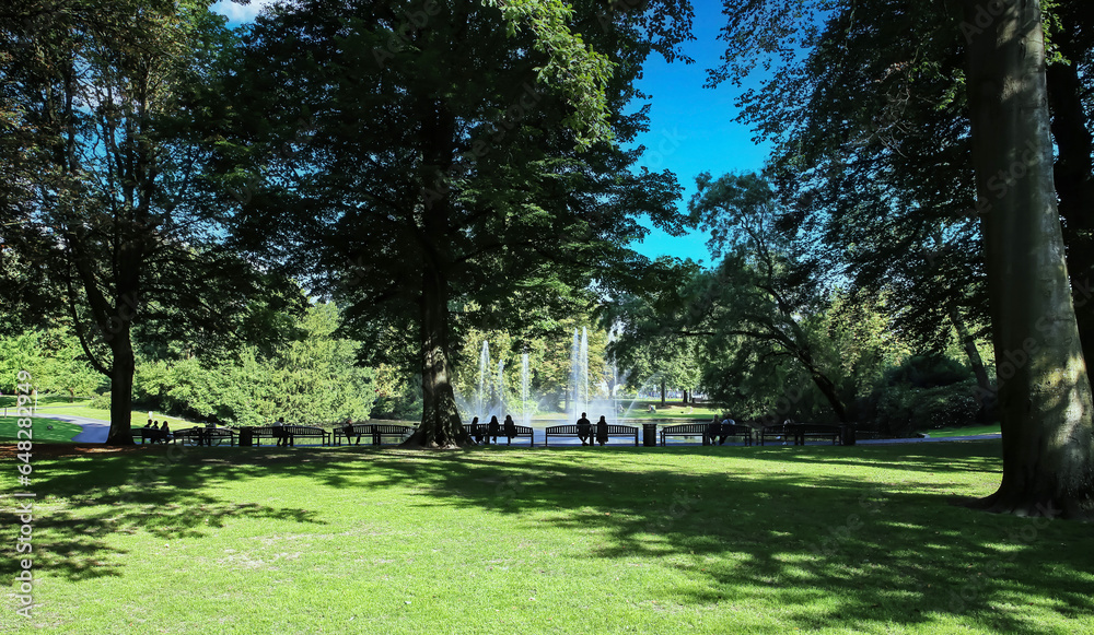 custom made wallpaper toronto digitalBreda, Netherlands - August 30. 2023:  Unrecognizable people sitting on benches in shade of trees at beautiful pond,  water fountain in green city park Valkenberg on sunny summer day