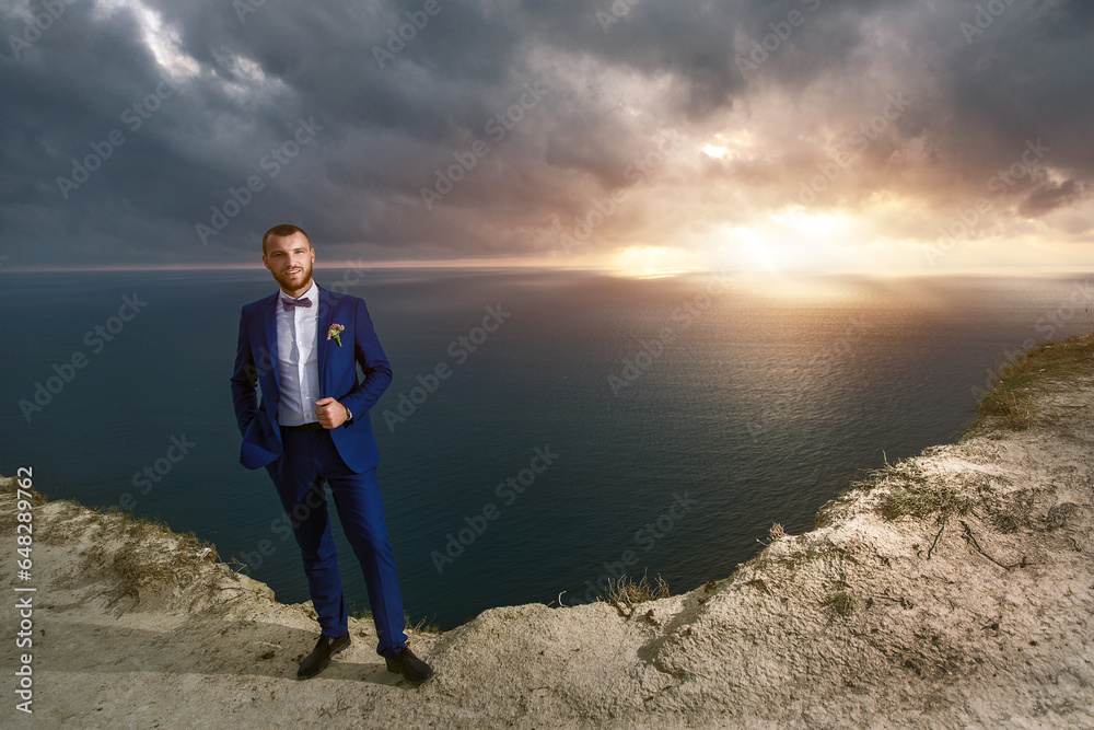 Fashionable groom in a businessman's suit stands on a mountain cliff overlooking the ocean, a man at a wedding photo shoot. Fashionable groom in a businessman's suit stands on a mountain cliff overlooking the ocean, a man at a wedding photo shoot.
