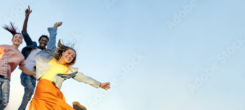 Happy multiracial young friends jump with raised arms isolated on blue sky background - Friendship concept - Copy space