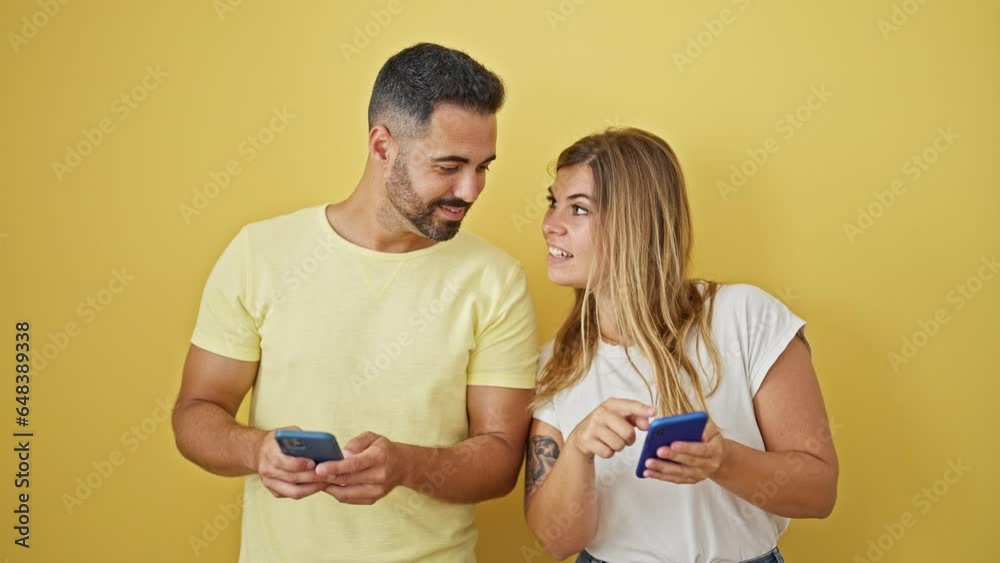 Man and woman couple smiling confident using smartphones over isolated yellow background Man and woman couple smiling confident using smartphones over isolated yellow background
