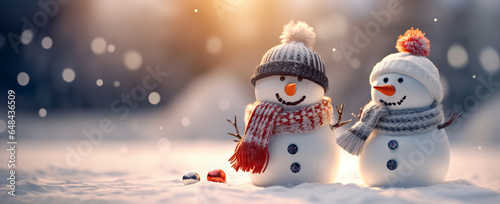 Two cute little snowmen are standing in a snowy landscape against a blurred background. They are smiling and there is enough negative space for copy space.