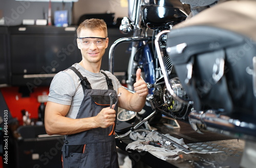 Motorcycle repair technician holds thumbs up while standing in workshop. High-quality service of transport equipment concept