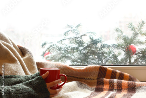 red mug in hands against the background of a window decorated with fir branches. cozy dreaming on winter holidays
