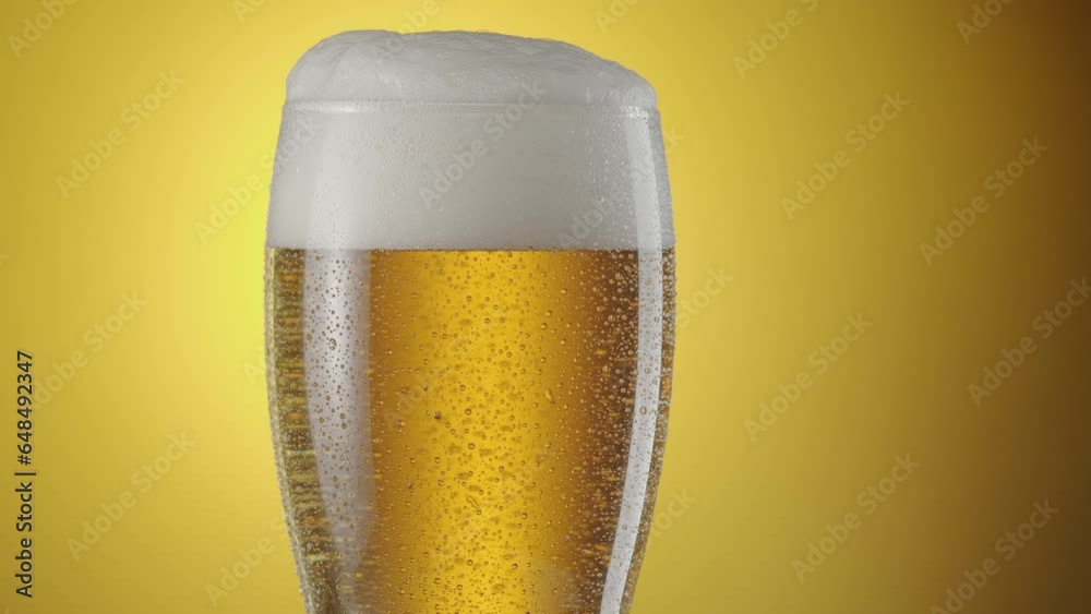 Camera slowly rises up along a beer glass with light beer. There are a lot of condensation drops on the glass, beer foam on top. Yellow background. Camera slowly rises up along a beer glass with light beer. There are a lot of condensation drops on the glass, beer foam on top. Yellow background.