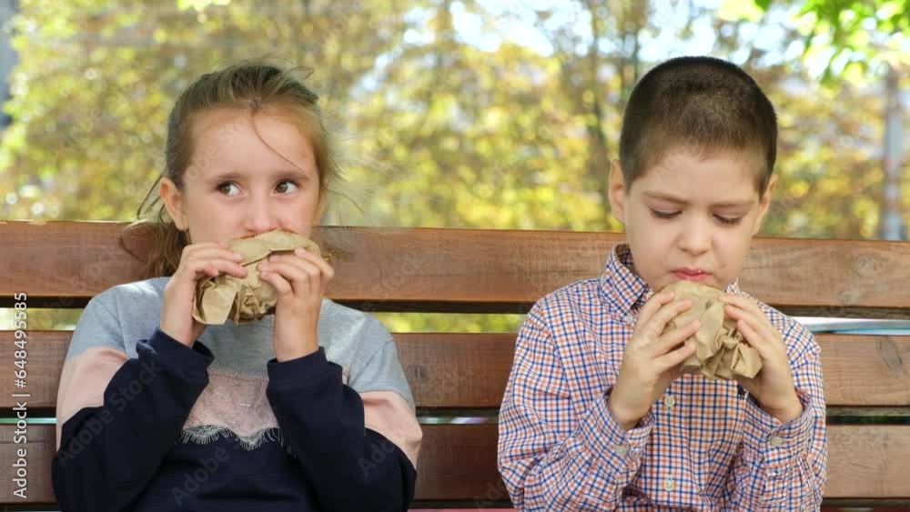 Small children, a boy and a girl eat burgers, street food sitting on a bench in the summer in the city. Small children, a boy and a girl eat burgers, street food sitting on a bench in the summer in the city.