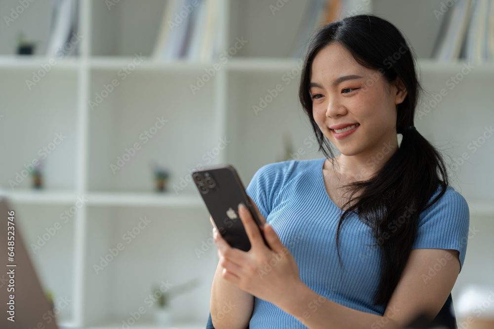 Asian woman sitting at work happily, smiling, delighted with business success feedback data from smartphone and data on laptop. while sitting on a desk in his office at home Asian woman sitting at work happily, smiling, delighted with business success feedback data from smartphone and data on laptop. while sitting on a desk in his office at home