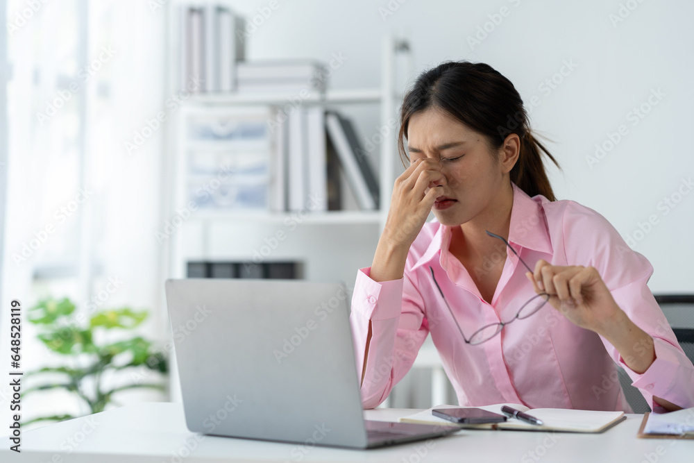 Asian businesswoman holding glasses thinking hard have pain around the eyes Tired of worrying about how to solve online document data by looking at the laptop screen in the office.