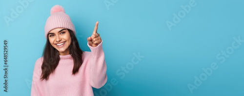 Portrait of a young smiling woman in knitted fashionable hat isolated on a flat blue background. Woman points her finger. Creative banner template for knitted hats store. 