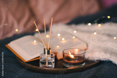 Home liquid fragrance in glass bottle and bamboo sticks with scented candle on wooden tray with open paper book close up over glow lights. Aromatherapy. Cozy home atmosphere.