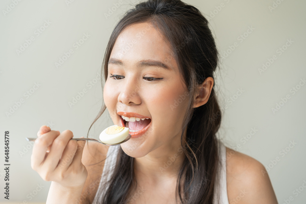 Protein in food, happy smiling asian young woman, girl in diet eat boiled egg, hand holding spoon put food in mouth with for breakfast in the morning at home. Health care, loss weight people concept. Protein in food, happy smiling asian young woman, girl in diet eat boiled egg, hand holding spoon put food in mouth with for breakfast in the morning at home. Health care, loss weight people concept.
