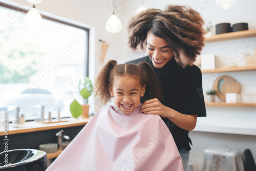 An African American girl laughs in a beauty salon while the hairdresser jokes with her, showcasing the friendship between a young girl and a hairdresser who share a fun and relaxed moment