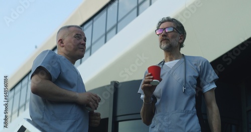 Two doctors in uniform stand near medical center entrance in the evening. Professional medics drink coffee during break and talk. Medical staff of hospital or modern clinic. Lifestyle and healthcare.