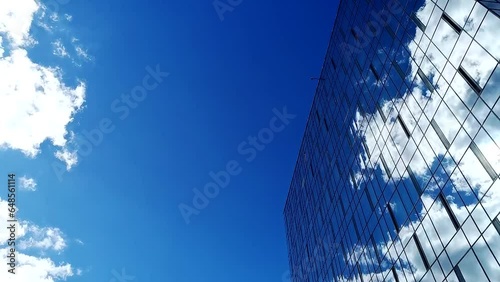 Time lapse of a blue summer sky with clouds reflecting off the glass facade of an office building. City office building with glass facade reflects white cumulus clouds floating in blue sky.