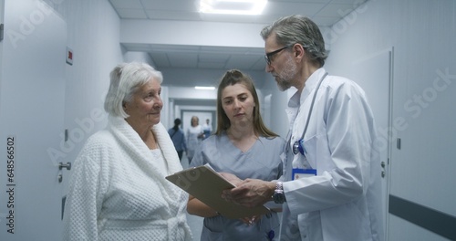 Mature doctor walks the clinic corridor, comes to colleague and elderly patient. Physician shows test results on papers to nurse and woman. Medical staff and patients in hospital hallway.