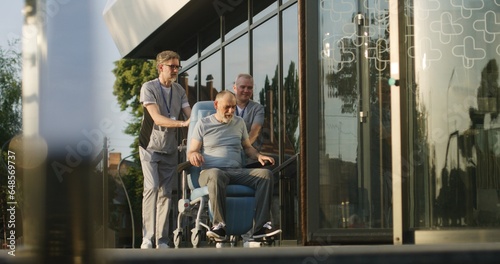 Two doctors in uniform take elderly man with disability to the clinic entrance on wheelchair for rehabilitation. Medical staff of hospital or modern medical center. Healthcare system. Slow motion.