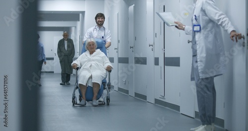 Doctor pushes wheelchair with female patient walking along clinic corridor. Mature medic opens door, another takes elderly woman to operating room. Medical staff and patients in hospital hallway. © Framestock