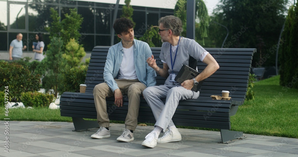 Mature doctor with tablet in hands sits with middle aged patient on bench and consults him. Man complains of leg pain to health care specialist. Medic examines clients leg and makes diagnosis outdoor. Mature doctor with tablet in hands sits with middle aged patient on bench and consults him. Man complains of leg pain to health care specialist. Medic examines clients leg and makes diagnosis outdoor.