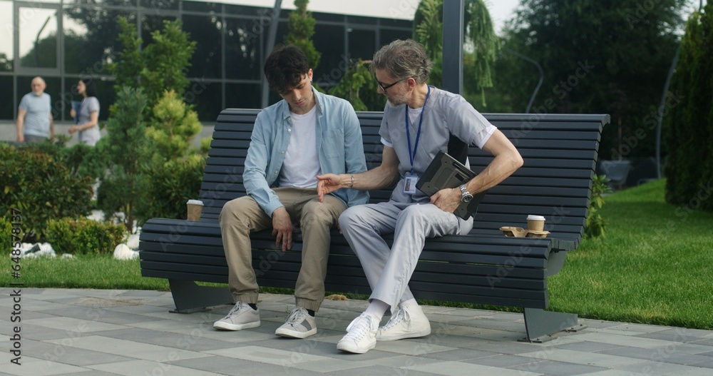 Mature doctor with tablet in hands sits with middle aged patient on bench and consults him. Man complains of leg pain to health care specialist. Medic examines clients leg and makes diagnosis outdoor. Mature doctor with tablet in hands sits with middle aged patient on bench and consults him. Man complains of leg pain to health care specialist. Medic examines clients leg and makes diagnosis outdoor.