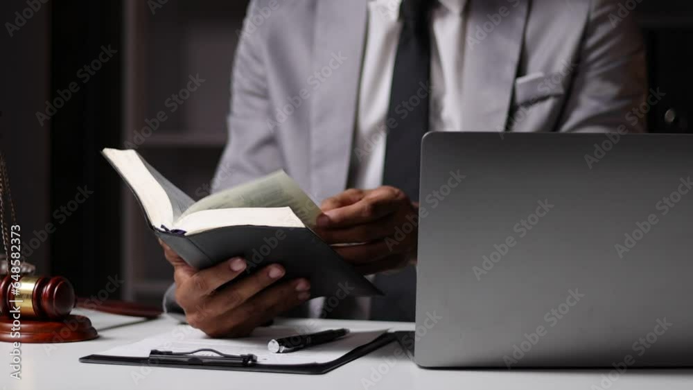 Lawyer reading law book and working on paperwork on laptop at desk in law office.