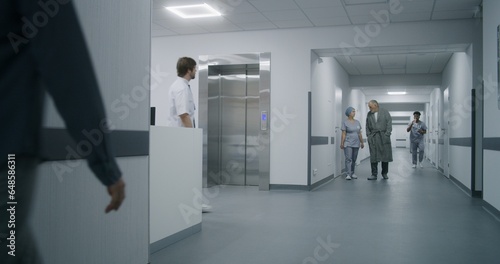 Nurse with old patient wait for elevator in medical center hallway. African American doctor walks down corridor with wheelchair. Caucasian medic goes to his patient. Medical staff at work in clinic.