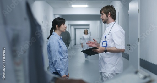 Male doctor stands in clinic corridor with female patient  uses digital tablet. Professional medic talks to adult woman  discusses medical tests results. Medical staff and patients in hospital hallway