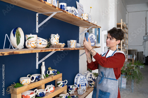 Charismatic Caucasian woman seller in souvenir shop takes pictures of goods at phone for posting on social networks. Successful girl store employee with ceramic tableware shoots video on smartphone photo