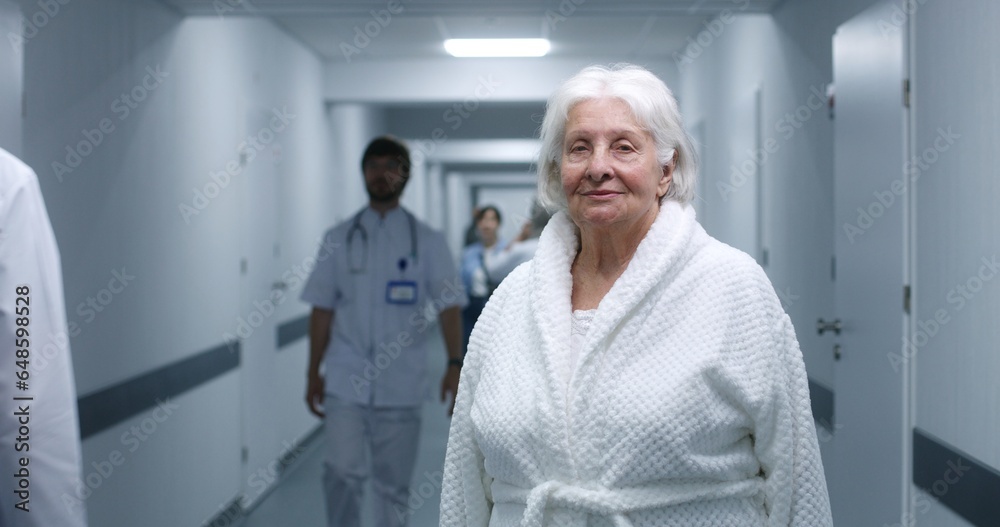 Obraz premium Senior woman stands in modern clinic corridor. Elderly female patient smiles and looks at camera. Medical staff, doctors and patients in the background in hospital or medical center hallway. Portrait.